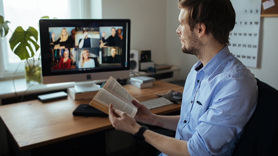 A man holds an open book while looking at a computer screen displaying six other people in a video chat