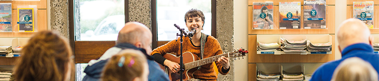 A person playing guitar and singing in a library with an audience watching
