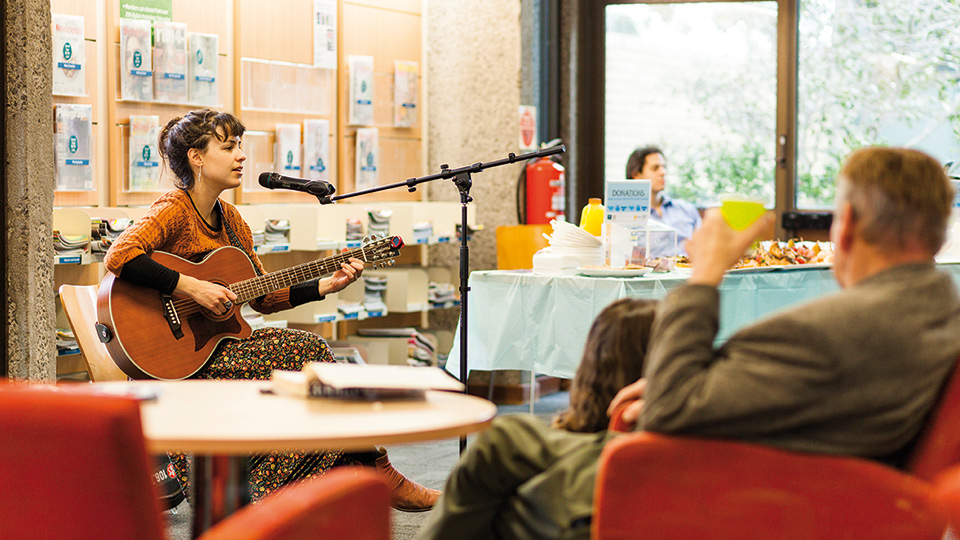 A woman in a library singing and playing the guitar with a man watching and seated on a red chair