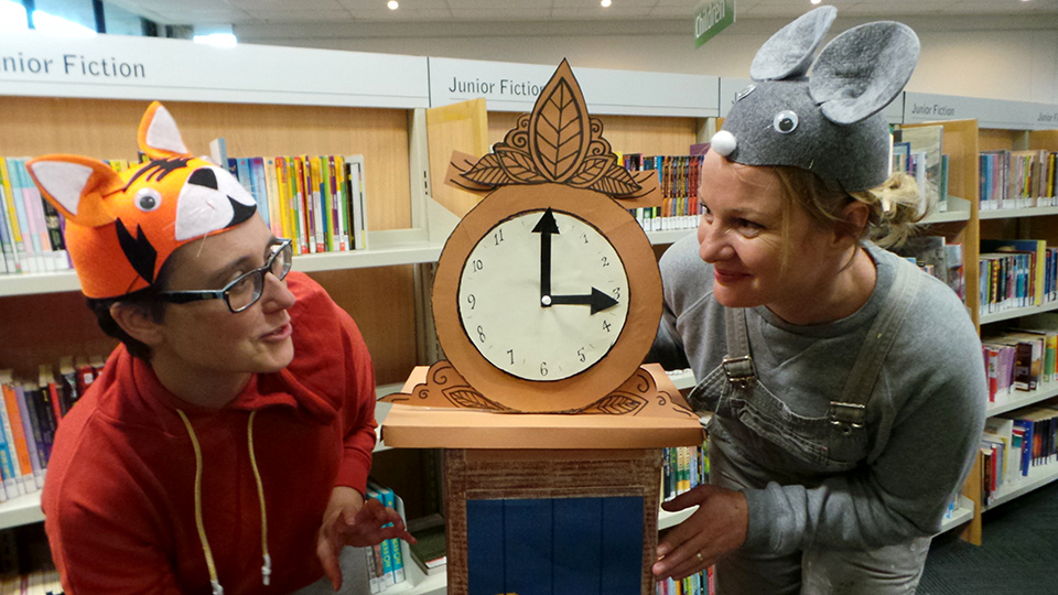 Two people stand on each side of a cardboard clock, wearing hats themed as cartoon-like cat and mouse