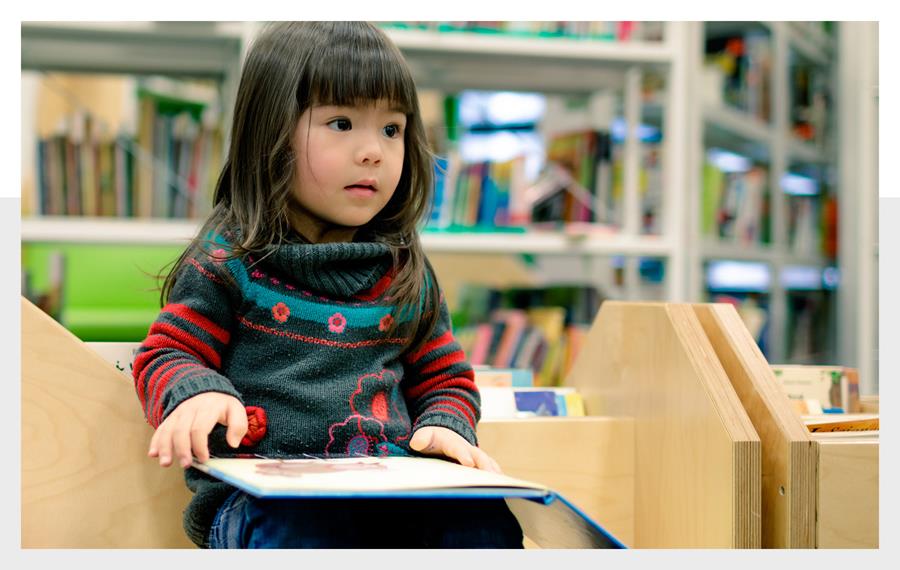 Photo of a young child sitting in a library. She has a picture book open on her lap and is looking at someone out of frame. She wears a grey knitted jumper with red and pink flowers. Behind her shelves of books are visible. 