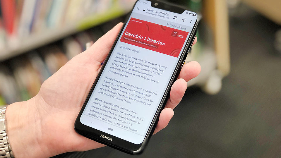 A hand holding a Nokia mobile phone displaying on the mobile phone screen an e newsletter from Darebin Libraries in the foreground.  In the background blurred bookshelves with books. 