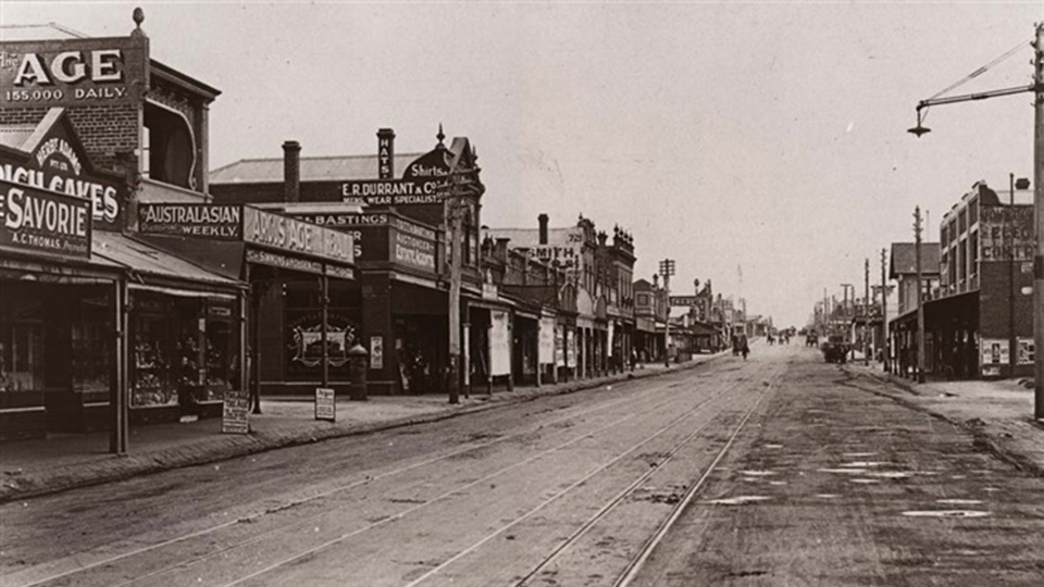 Photo cypher tone image of  turn of the century 1900s of a high street with shops and tram lines in the foreground.  Background are people walking and crossing the road. Shops in the foregrounds have advertising boards and signage on the side of the brick buildings