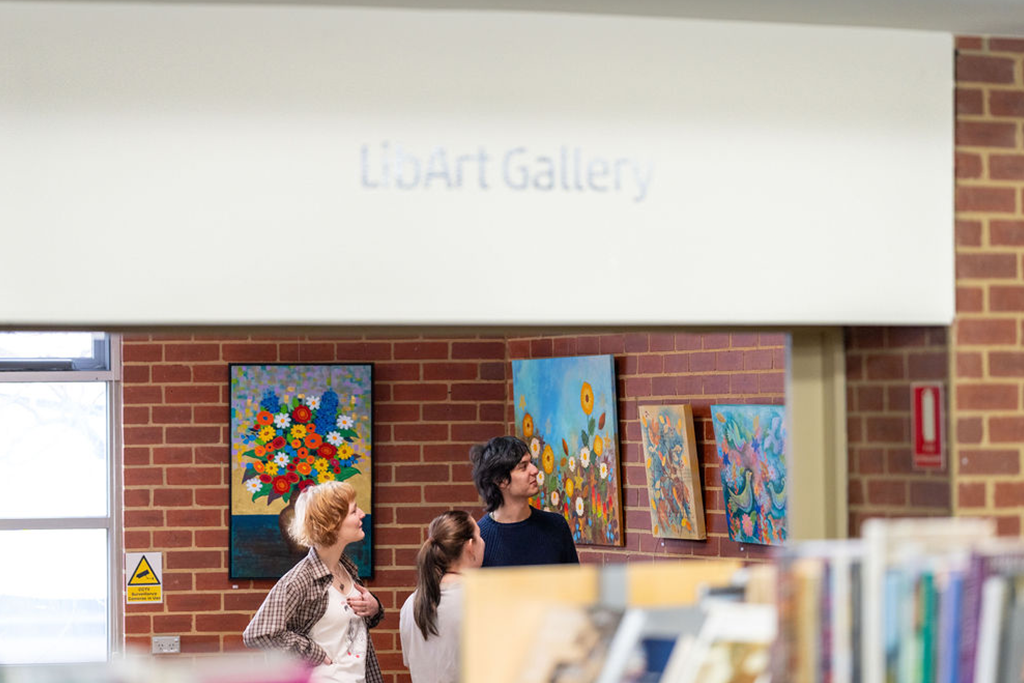 Photo of a room with four colourful paintings of flowers hanging on brick walls. Three young people are looking at one of the paintings and smiling. In the foreground a sign says LibArt Gallery and a bookshelf is visible but out of focus. 