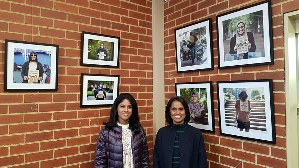 Two ladies smiling and standing in front of an inside corner brick wall.  Behind  them photographs on the wall of diverse people. 