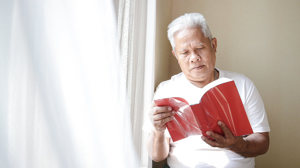 An aged man standing against a wall reading a red book