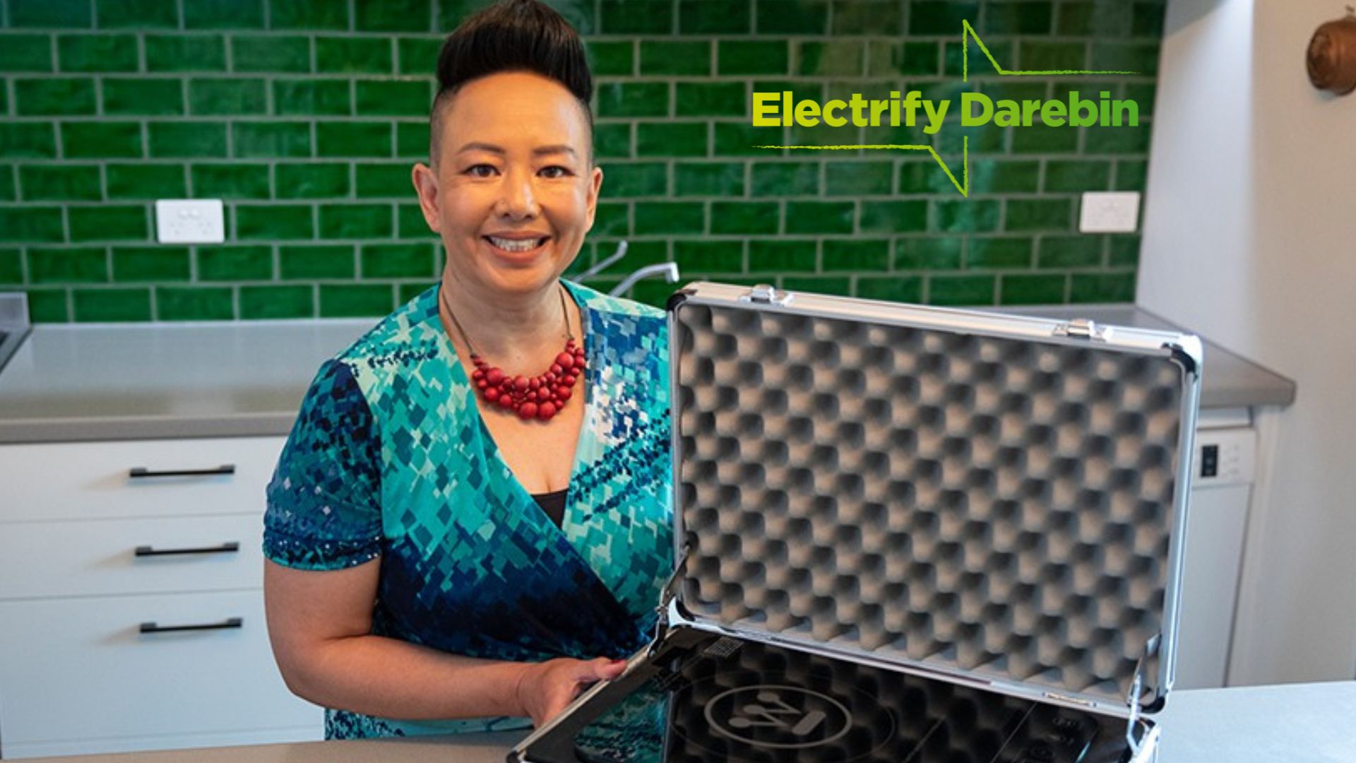Image: Photo of a smiling person with short dark hair and chunky red necklace standing in a kitchen and holding an open case containing an induction cooktop.