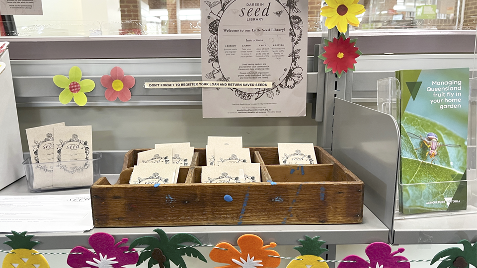 Photo of a table with a collection of seeds in packets with Darebin Seed Library branding. The table is decorated with flowers, information signs about the seed library, and gardening brochures.