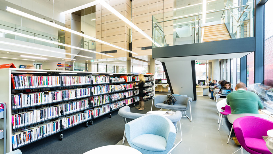 Inside of  modern library with high ceiling light and spaceous. Foreground on the right hand side are people sitting on chairs reading and working on computer. To the left are book shelves and a lady standing near a book shelf. Staircase in background on right hand side.