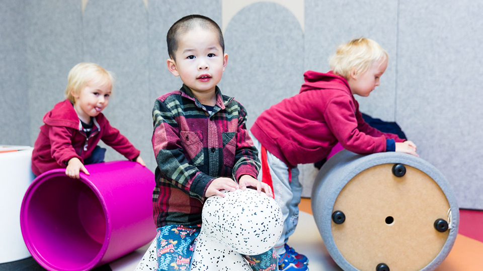 Three children indoors; one playing with a toy in foreground, the others pushing large toy cylinders in background
