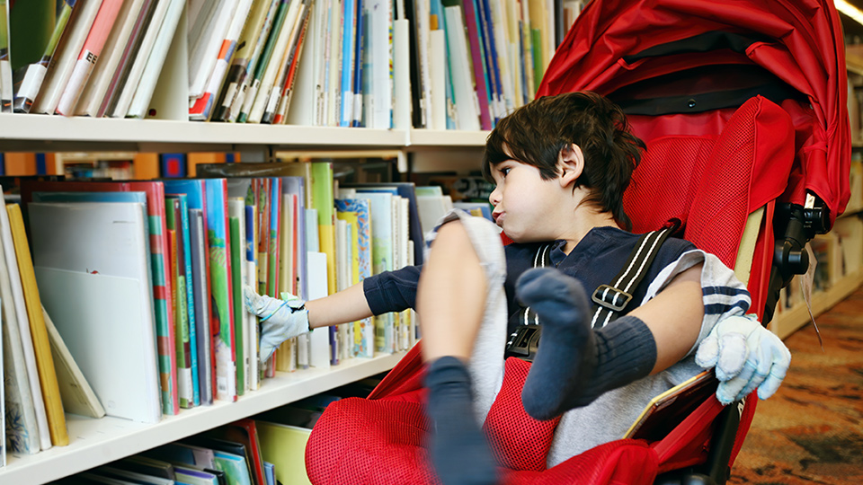 Child strapped into a red stroller and touching with one hand children's  books on a book shelf.