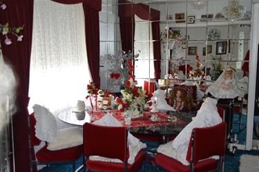Margaret's Simmons Dining Room table is placed before a mirror wall. There are porcelain dolls sitting at the table. The space is decorated with white late and red velvet curtains and chairs.