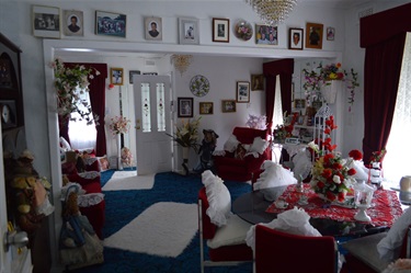 Margaret's Simmons Dining room features red velvet furniture and blue carpet. The room is decorated with porcelain dolls, white doilies, while pillows and many family portraits.