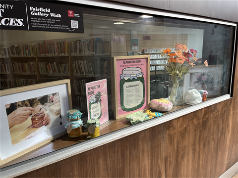 Photo of a gallery window displaying jars of relish, knitted mittens and hats, a vase with orange flowers, and a framed graphic of a jar.