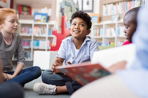 Photo of three school-aged children in casual clothes. They are sitting on the floor and chatting. In the background bookshelves are visible. 