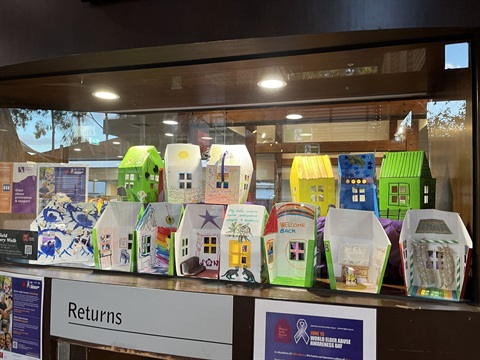 Photograph of thirteen small paper houses on display in a glass cabinet. The houses have been coloured in and decorated with drawings, pictures, cutouts, ribbons, and have messages of welcome written on them.