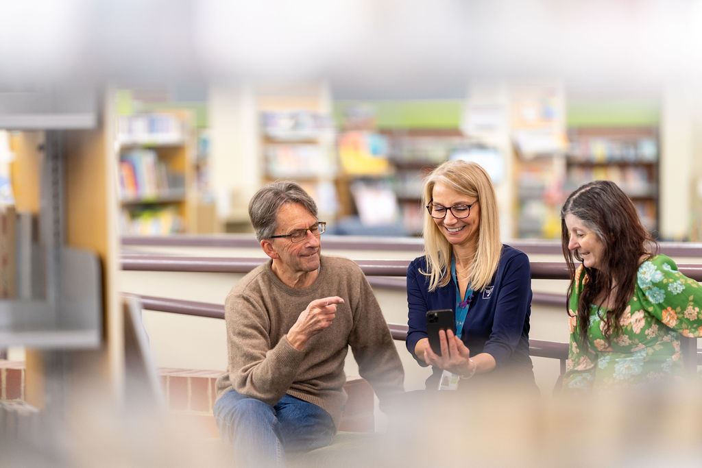 Photo of three people sitting in a library. The middle person is in Darebin Libraries uniform and is showing the other two something on a phone. 
